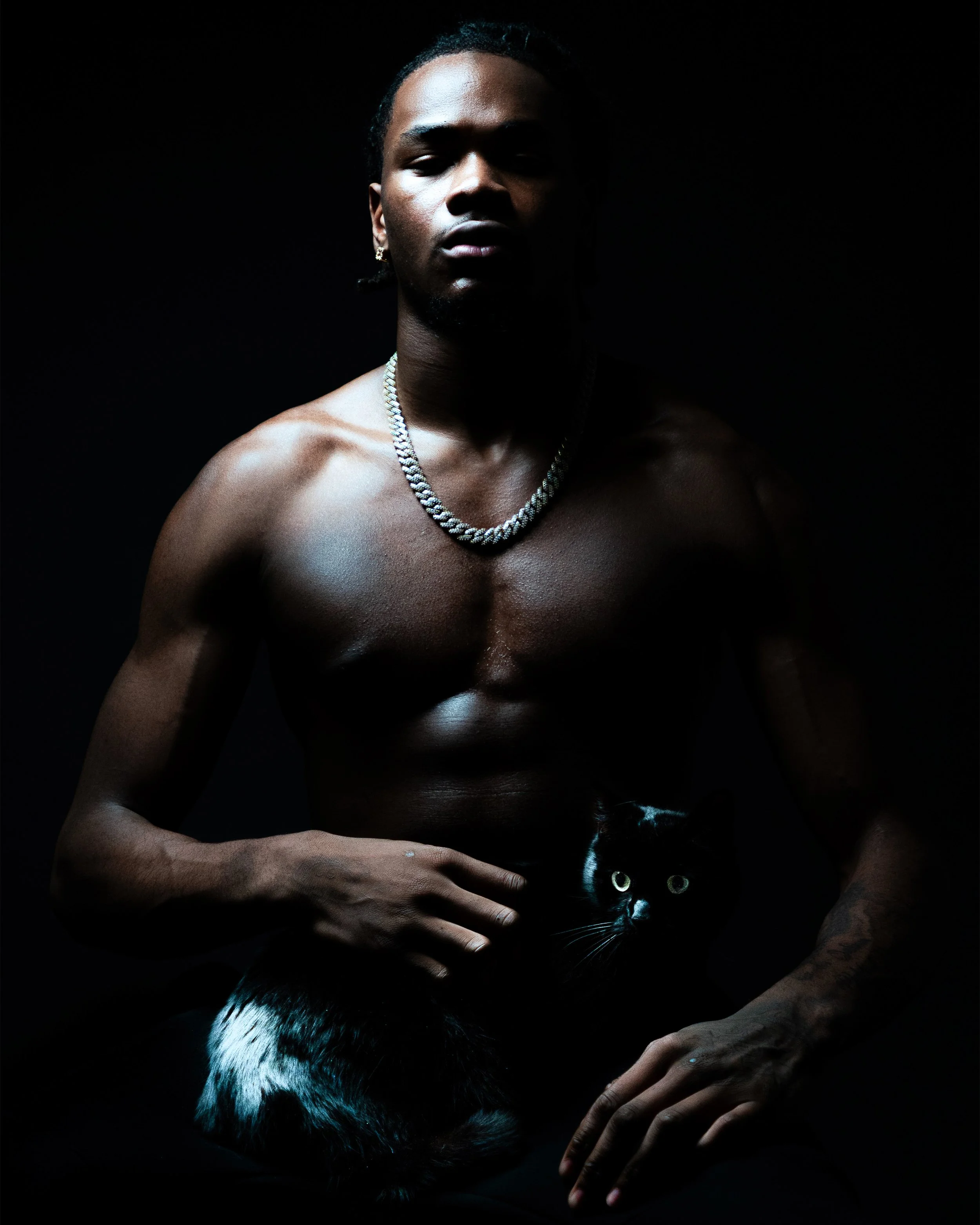 Un homme nu avec un collier en chaîne, tenant un chat noir avec des taches blanches, dans un environnement sombre.