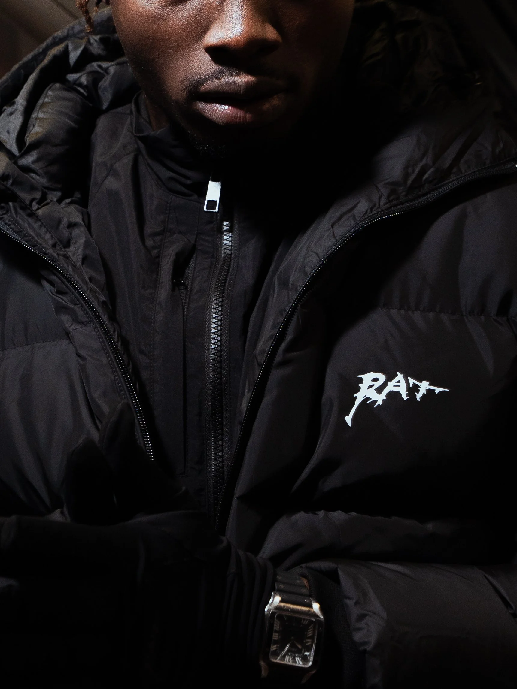 Homme portant une veste noire avec le logo RAY, montre argentée, la moitié du visage visible, fond sombre.