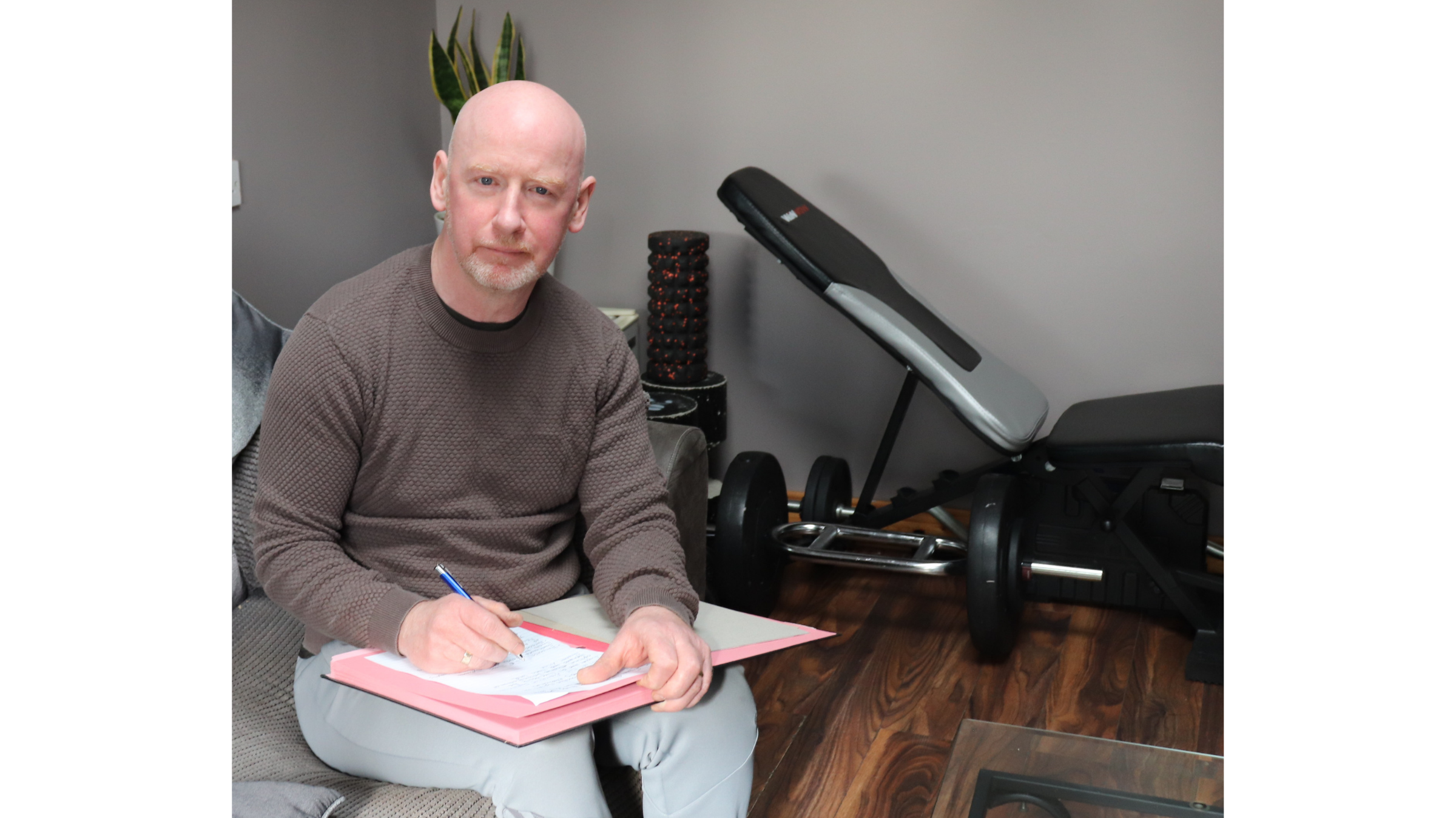 A bald man with a beard sitting on a chair in a room, writing in a red folder with a pen. There is a plant and exercise equipment behind him.