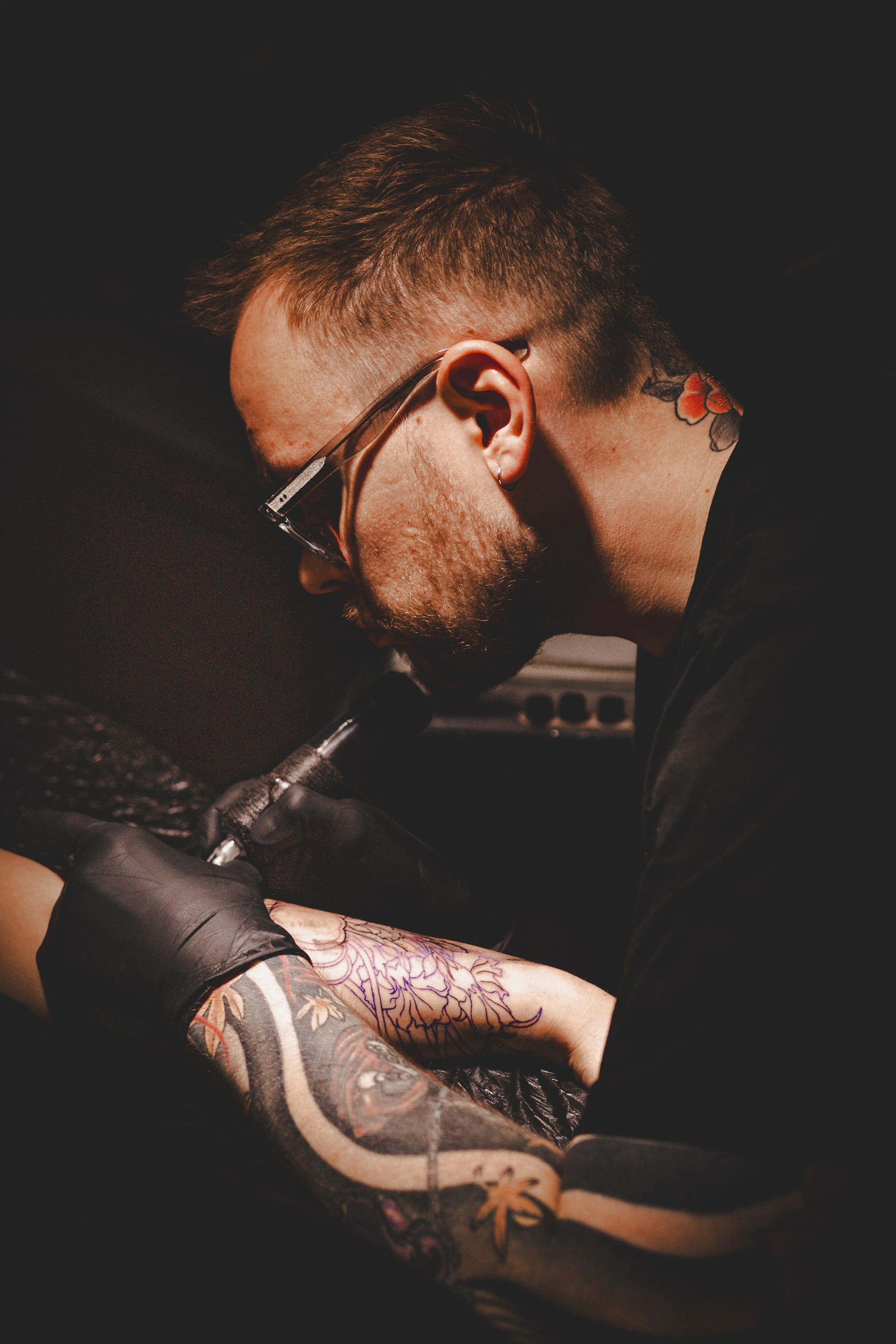 Ein Mann mit Tattoos und Brille beim Tätowieren eines Armes, getragenes schwarzes T-Shirt, dunkler Hintergrund.