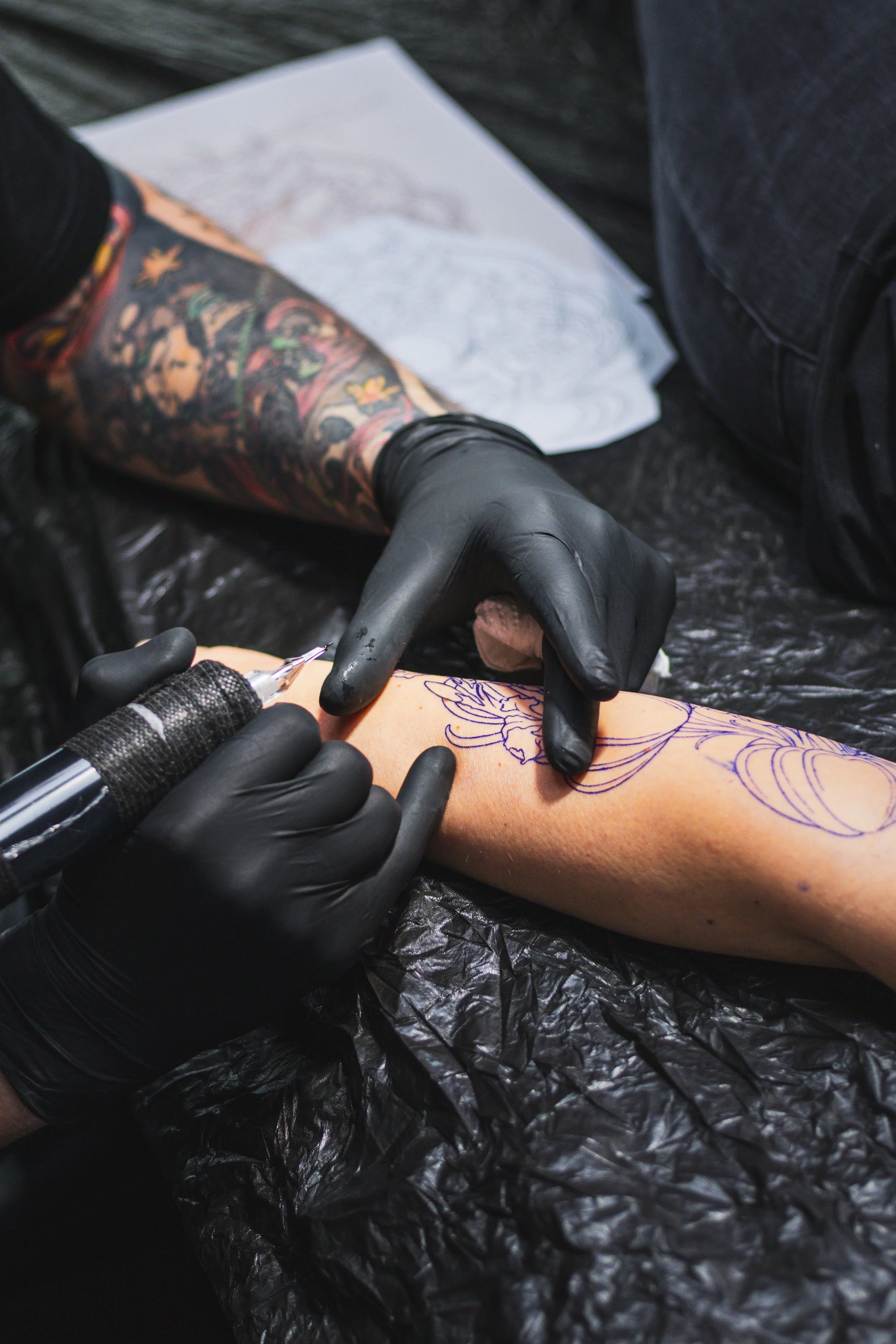 Tattoo-Künstler tätowiert eine Person am Arm mit einer Vorlage und einem Tätowierstift, während er schwarze Handschuhe trägt.