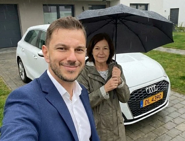 Un homme en costume bleu prend une selfie avec une femme tenant un parapluie noir devant une voiture blanche, sous une pluie légère, dans une cour résidentielle.