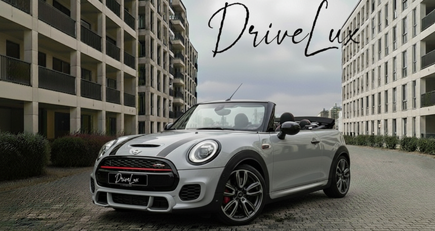 Une voiture décapotable grise, une Mini Cooper, garée entre deux bâtiments modernes, avec un texte en arrière-plan qui dit 'DriveLuxe'.