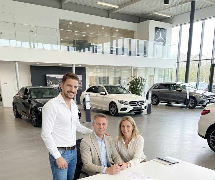 Trois personnes souriantes dans un concessionnaire automobile, avec des voitures Mercedes-Benz en arrière-plan.