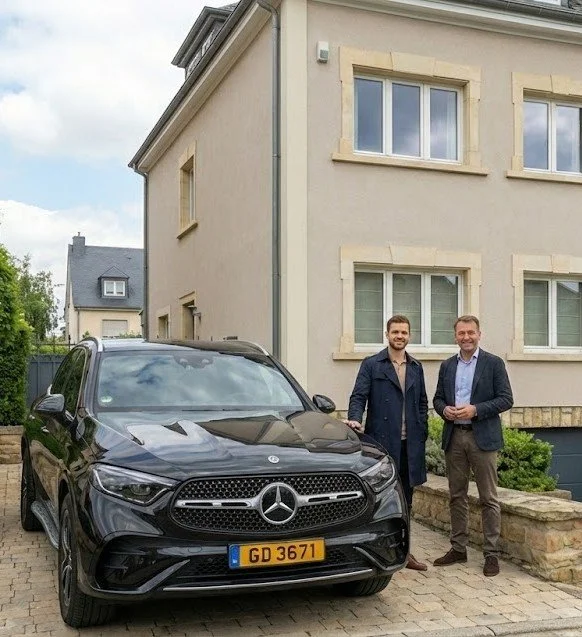 Deux hommes souriants se tiennent à côté d'une voiture Mercedes noire stationnée devant une maison. La maison est beige avec plusieurs fenêtres et un mur en pierre devant. Les hommes portent des costumes et semblent être en train de poser pour la photo.