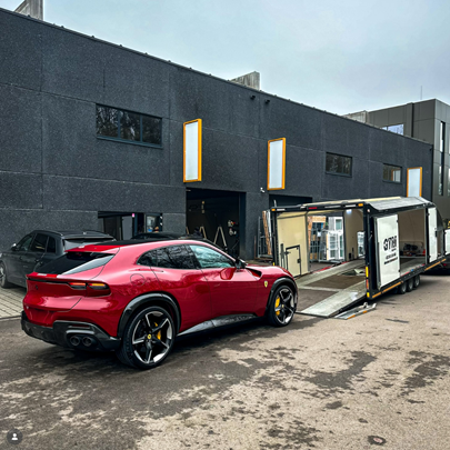 Voiture rouge de luxe stationnée devant un bâtiment moderne, avec une remorque ou un stand de vente de voitures à côté.