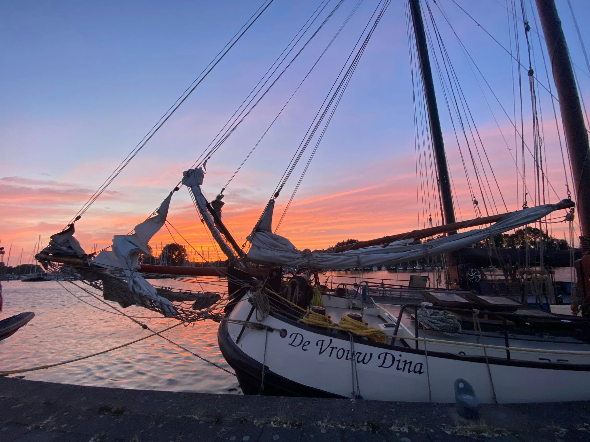 Zeil-tjalk genaamd 'De Vrouw Dina' afgemeerd bij zonsondergang met zijlzeilen omhoog en kleurrijke lucht op de achtergrond in een havenomgeving.