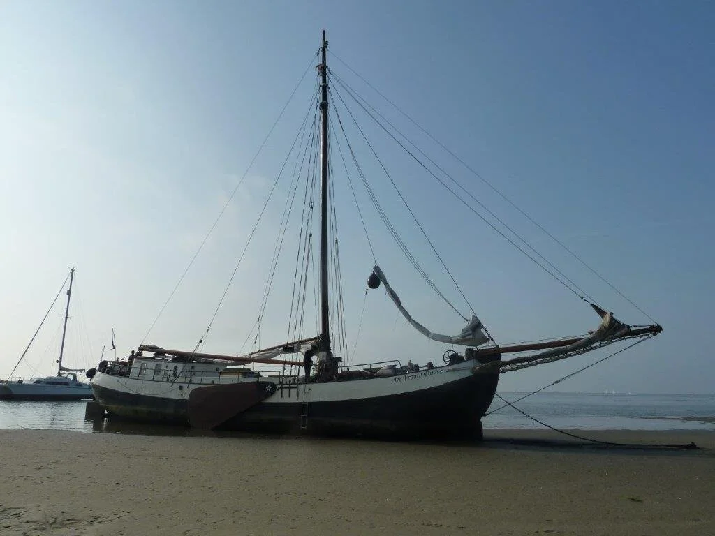 Een traditionele zeilboot ligt droog op het strand, met de mast rechtop en zonder zeilen. De achtergrond toont een andere boot op het water.