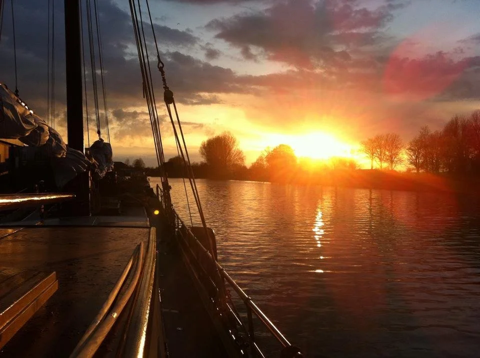 Zonsondergang over een meer met silhouetten van bomen en dekken van een zeilboot op de voorgrond.