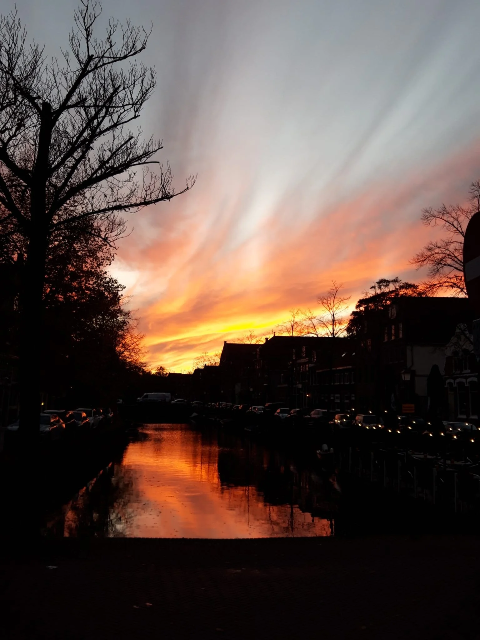 Silhouetten van gebouwen en bomen langs een kanaal bij zonsondergang in Nederland, met een dramatisch gekleurde lucht en reflecties in het water.