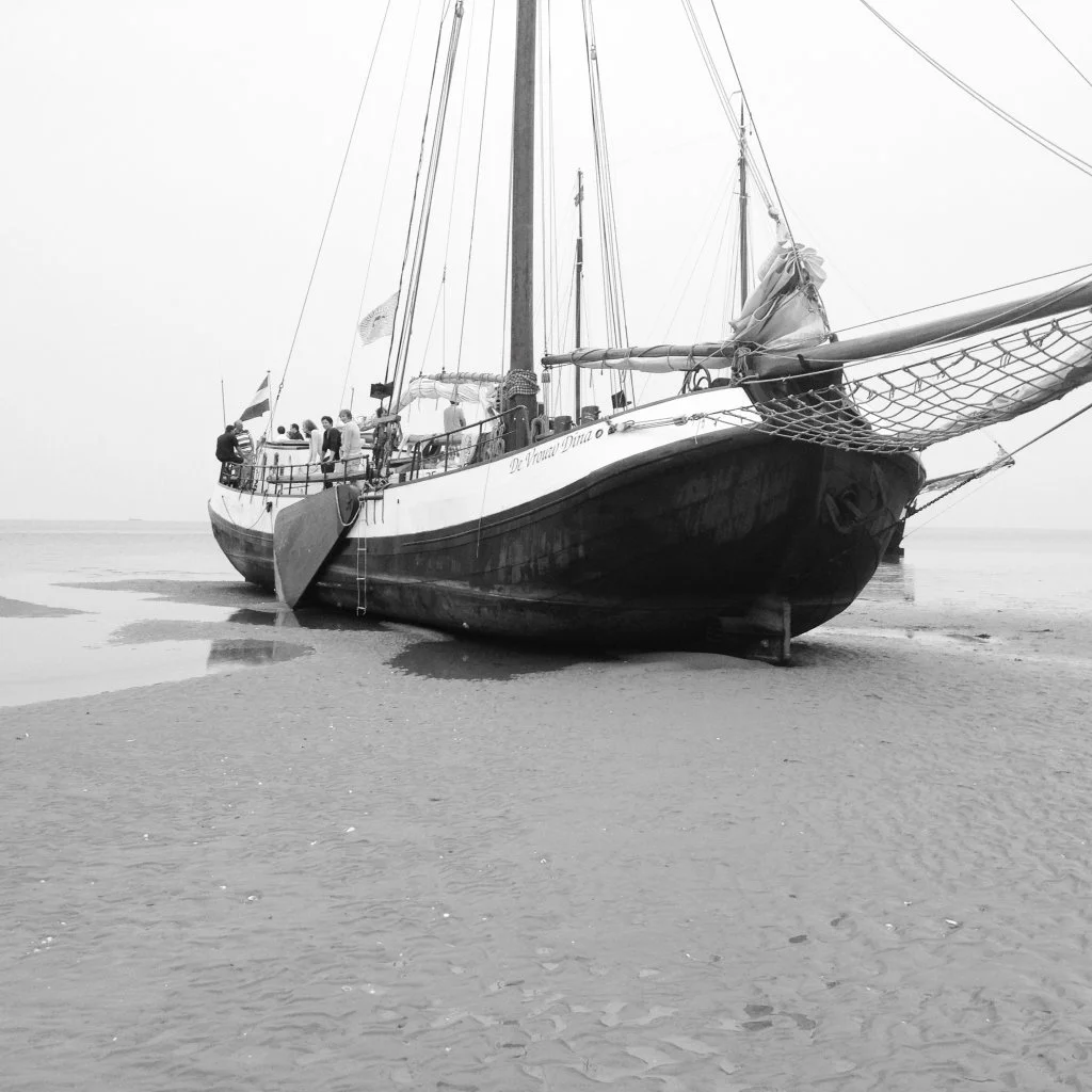 Een zeilschip op het droge bij laag water met mensen aan boord. Het schip ligt op zand, onder een bewolkte lucht in zwart-wit weergegeven.
