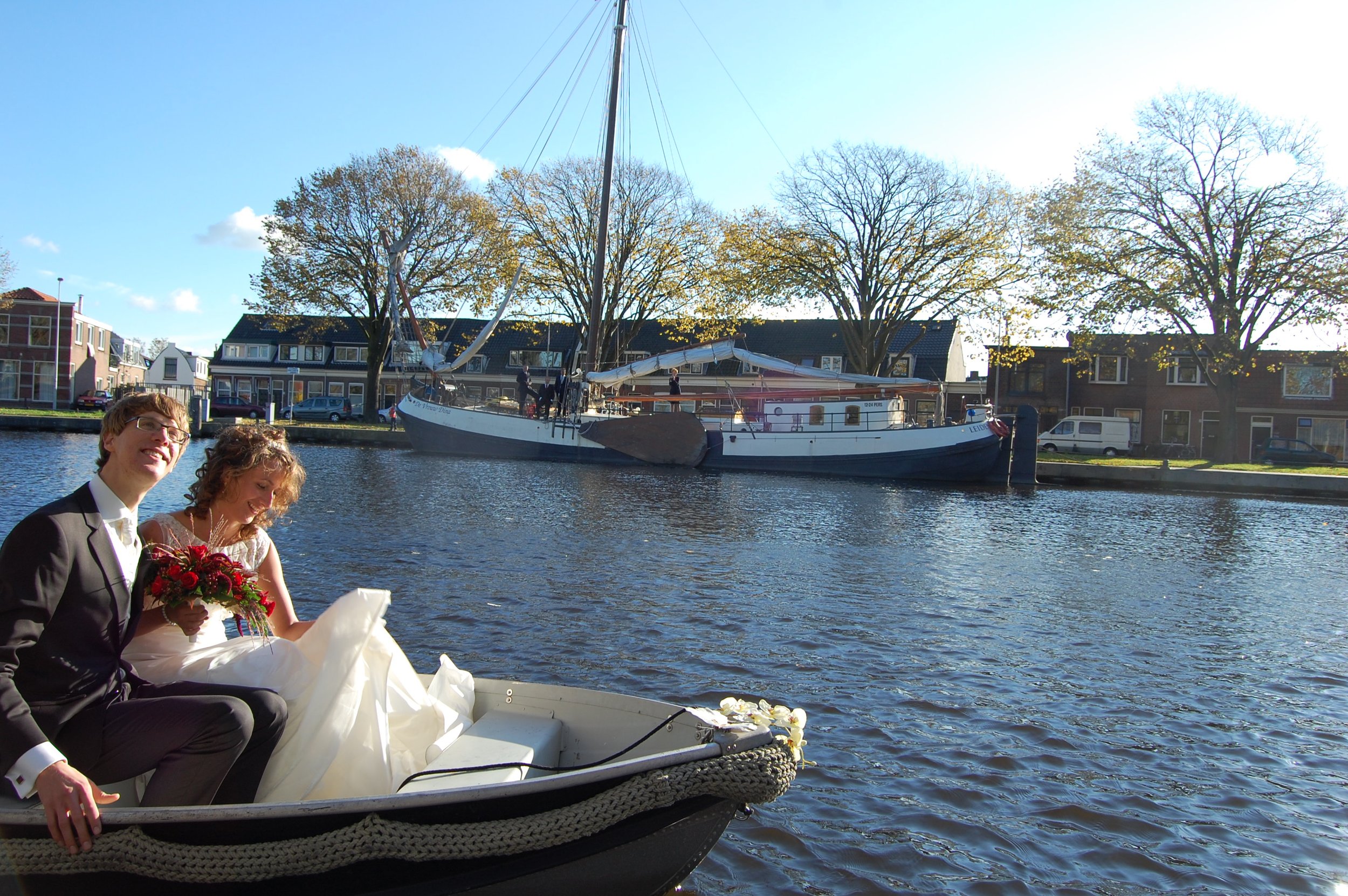Bruidspaar op een boot op de rivier met een traditionele zeilboot en gebouwen op de achtergrond.