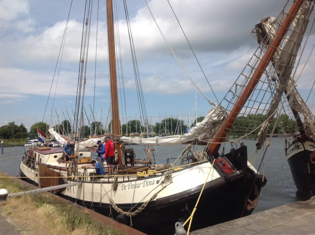 Traditionele Nederlandse zeilboot aangemeerd in een haven, met mensen aan boord en een Nederlandse vlag. Bomen en andere boten op de achtergrond.