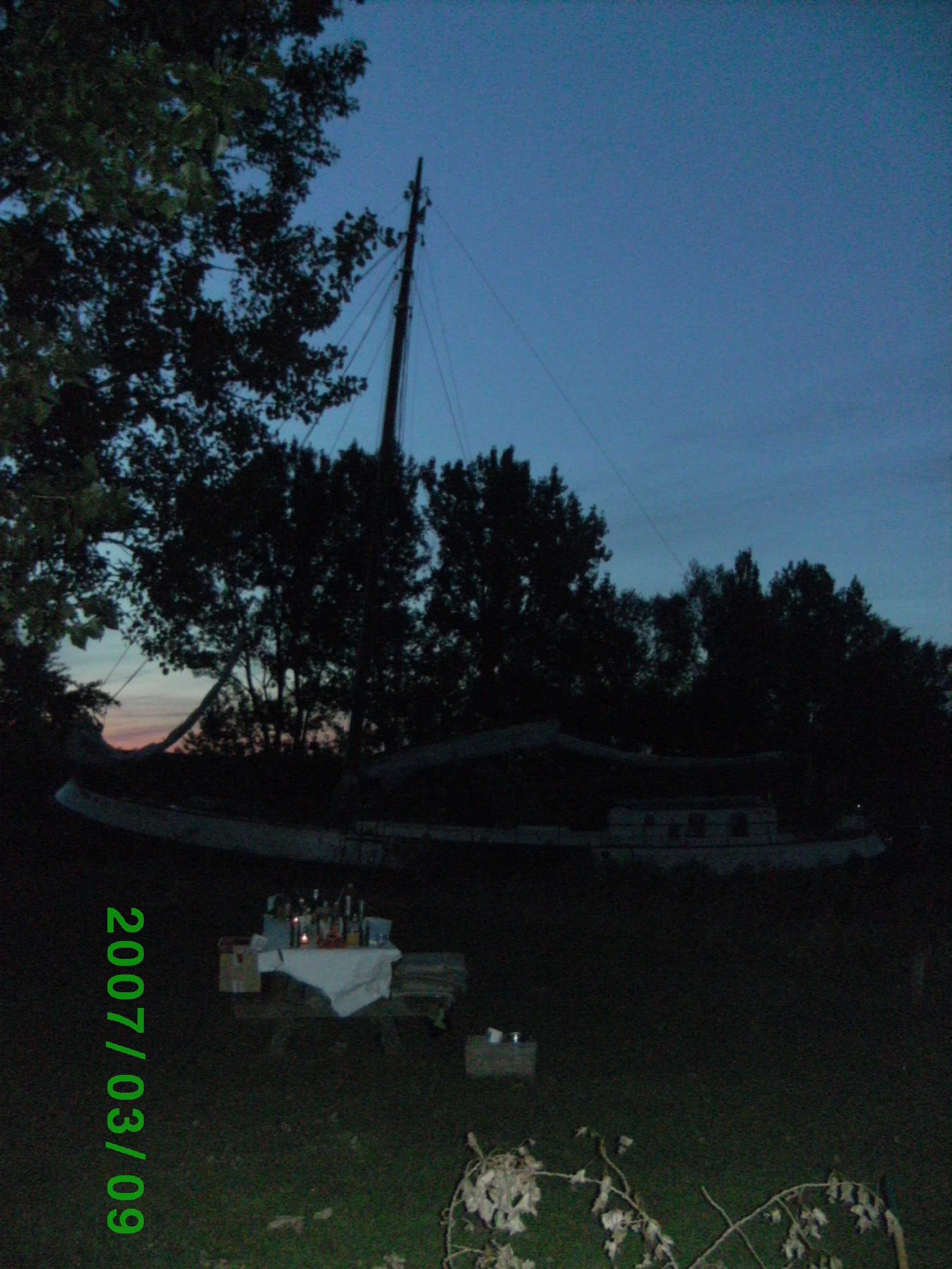 Schip op de achtergrond bij avondschemering, met een tafel bedekt met een wit tafelkleed en flessen op de voorgrond.