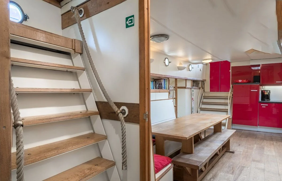 Interieur van een woonboot met houten trappen, eettafel, banken, rode keukenkasten en boekenplanken.