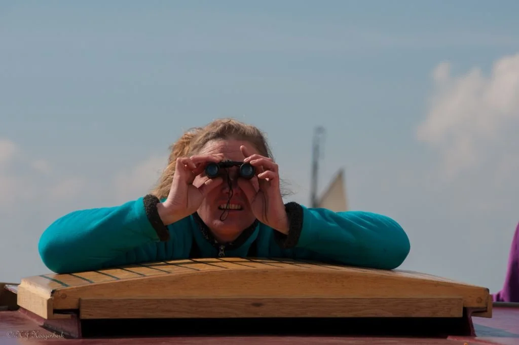 Vrouw met verrekijker kijkt uit vanaf een houten object tegen een blauwe hemel.