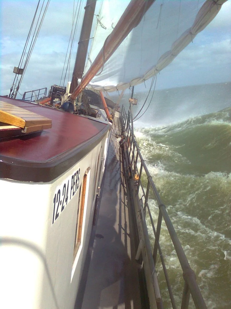 Zeilboot op zee, golven slaan tegen de romp, zeilen in de wind, blauwe lucht.