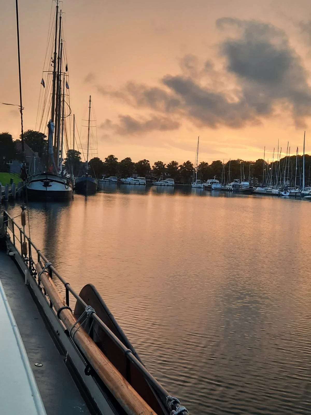 Zeilboten in een haven bij zonsondergang, met een rustige wateroppervlakte en een bewolkte lucht.