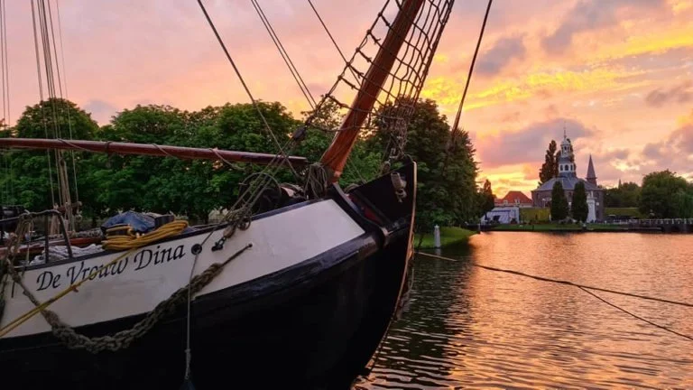 Een zeilboot genaamd 'De Vrouwe Dina' ligt aangemeerd bij de oever van een rivier tijdens een kleurrijke zonsondergang, met een kerk en bomen op de achtergrond.