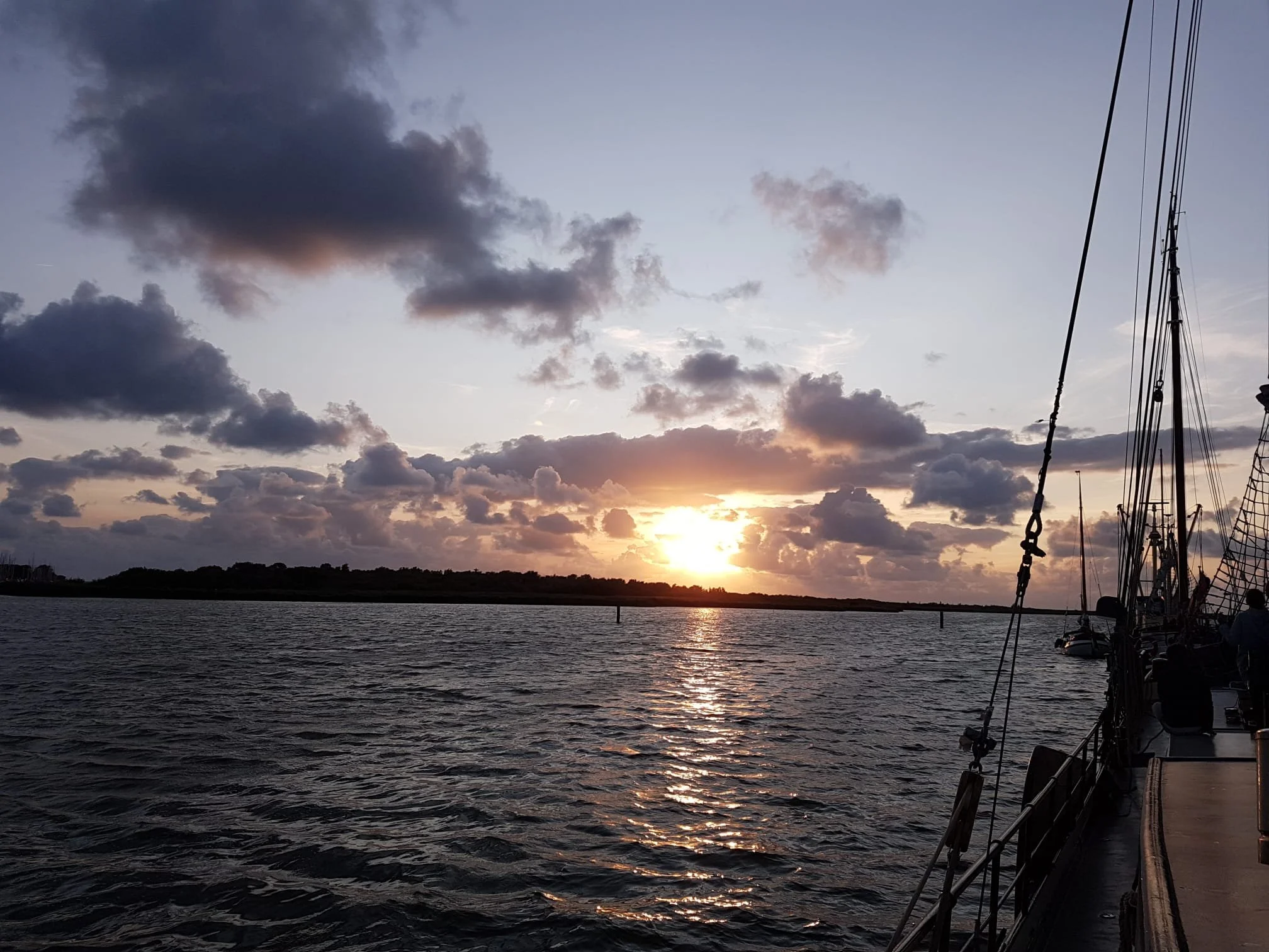 Zonsondergang boven een zee met een silhouet van een zeilboot op de voorgrond en wolken aan de hemel.