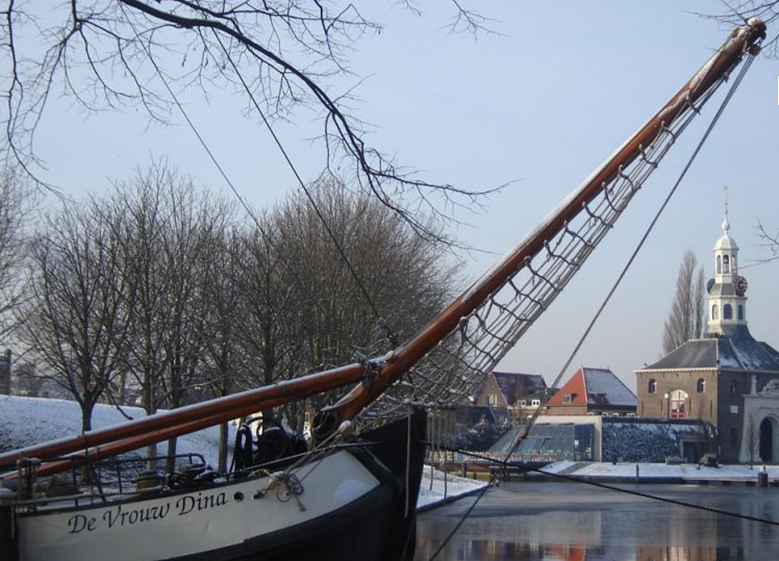 Traditioneel zeilschip 'De Vrouw Dina' aangemeerd in een winterse haven, met bomen op de achtergrond en een kerk naast gebouwen.