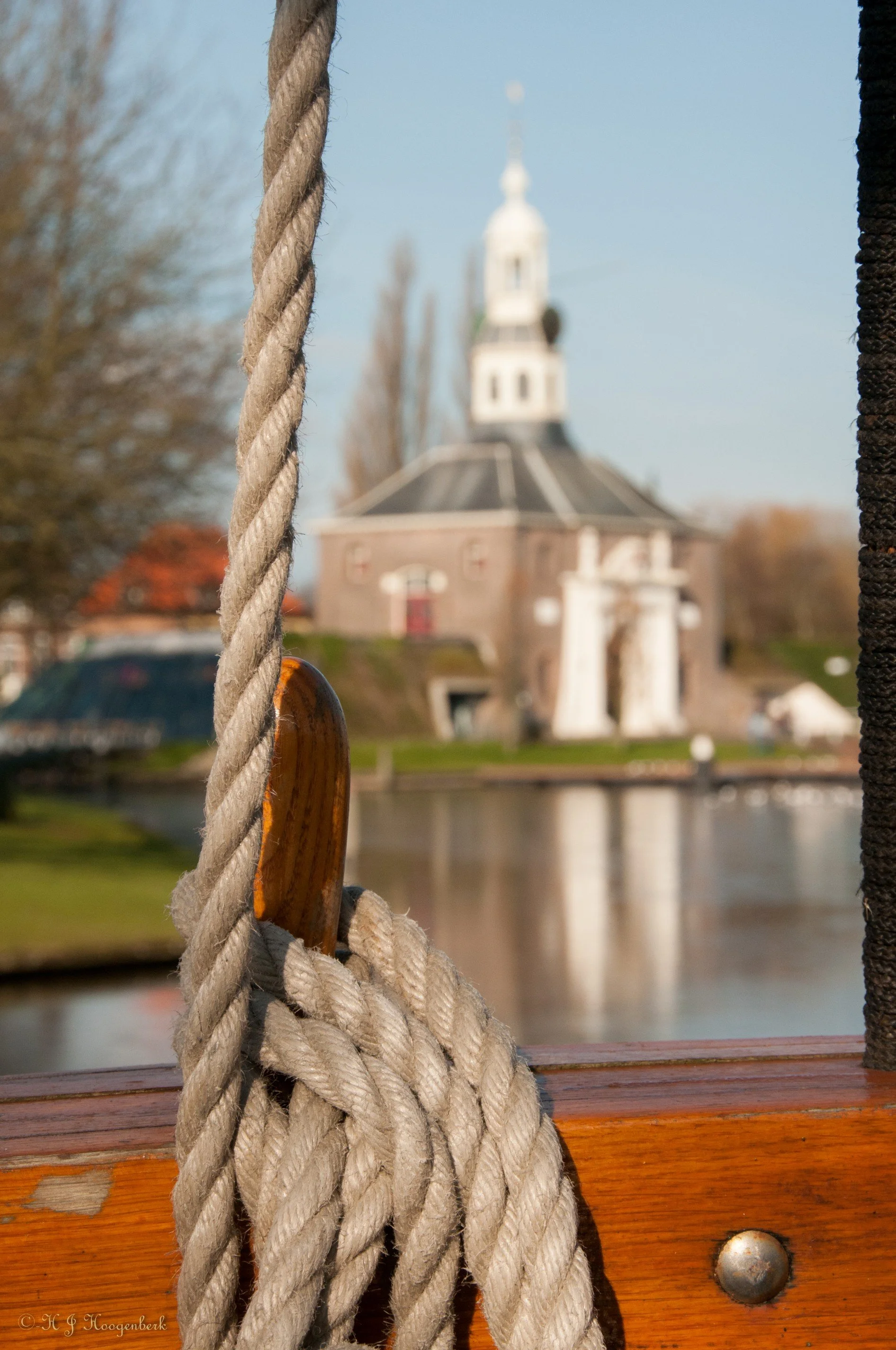 Touw op een houten reling met historische toren op de achtergrond, weerspiegeld in het water.