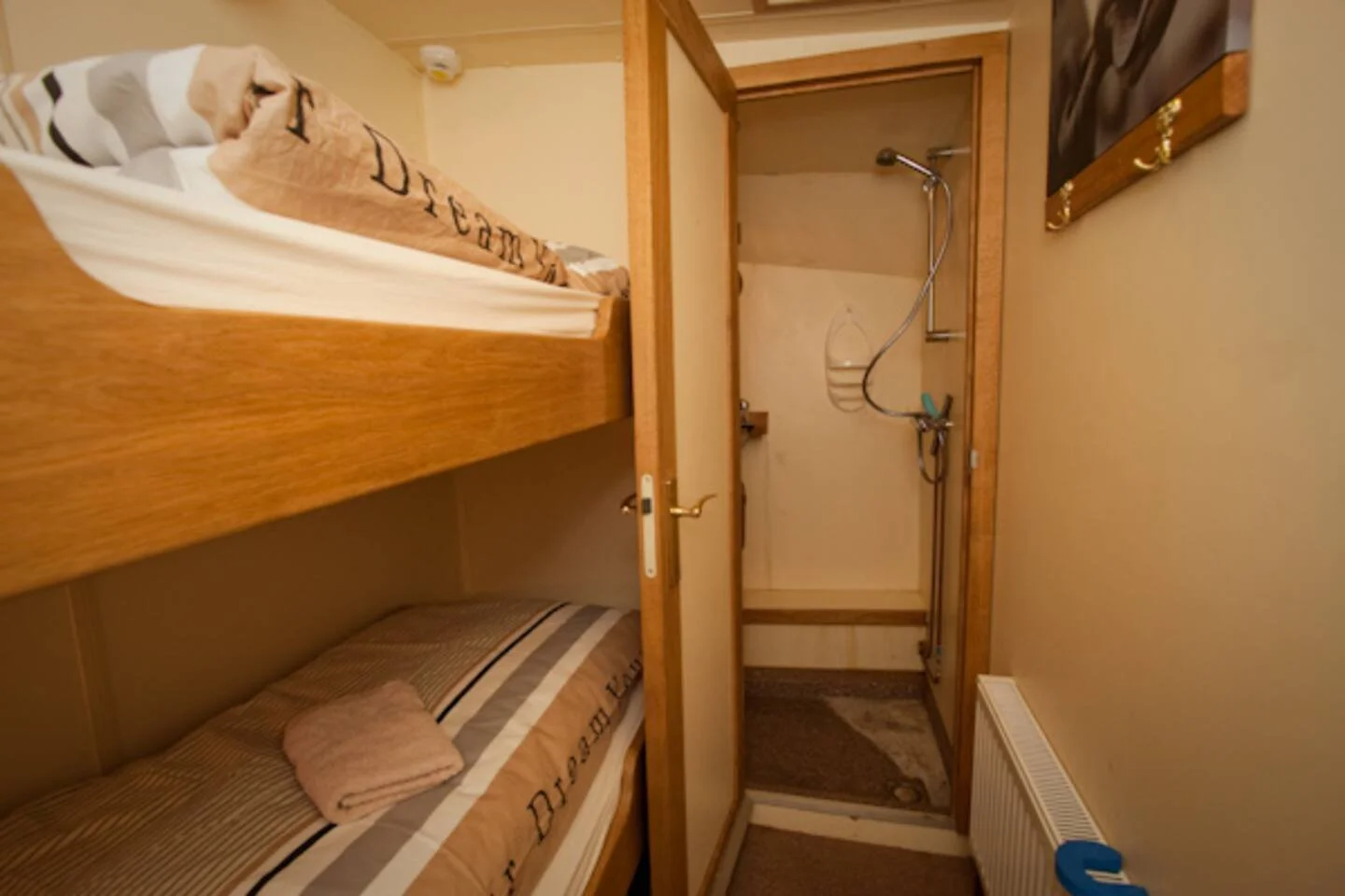 Kleine slaapkamer met stapelbed, handdoek op bed, open deur naar douche, radiator en muurdecoratie.