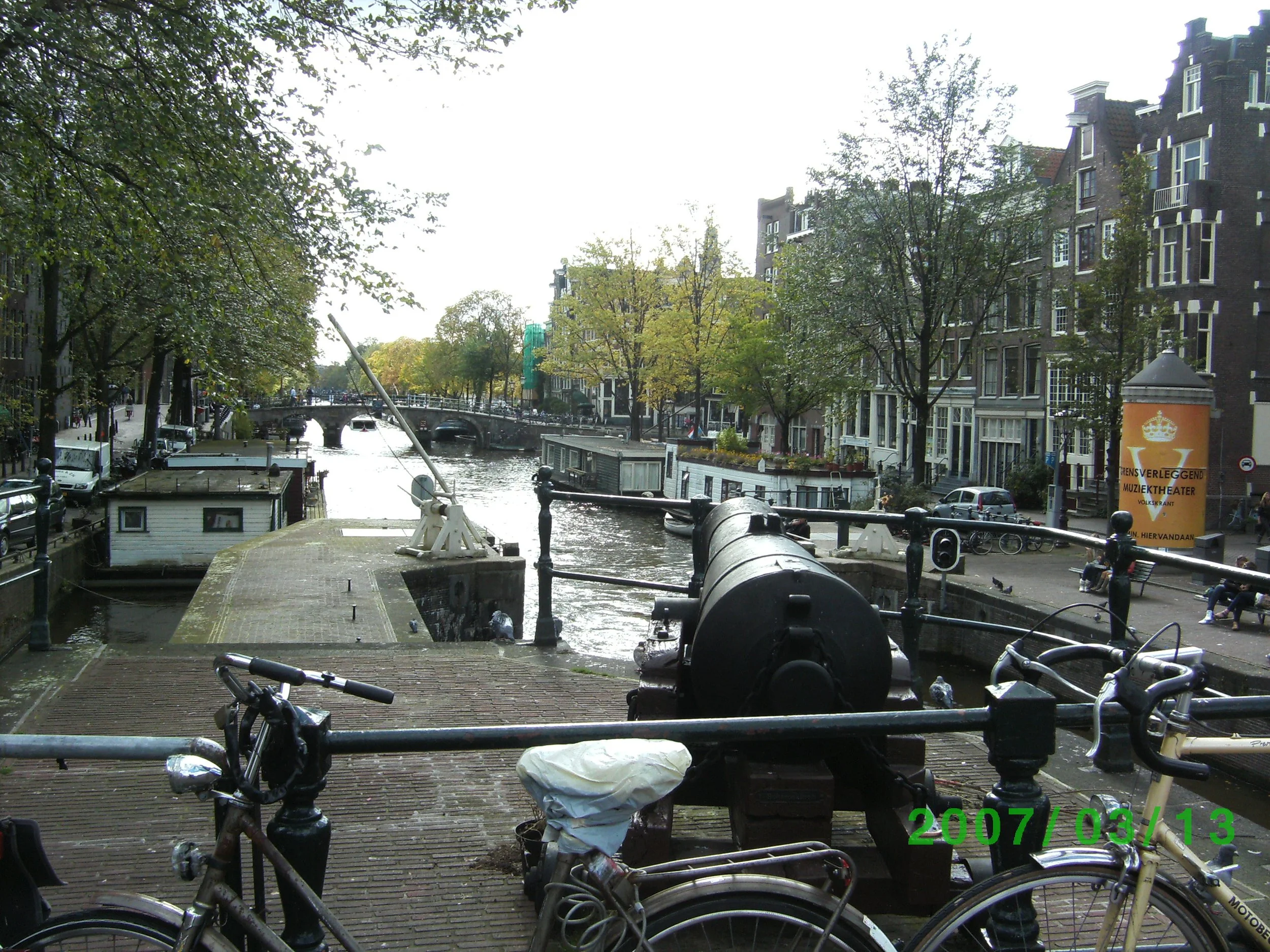Uitzicht op een Amsterdamse gracht met fietsen op de voorgrond, een oud kanon en woonboten langs het kanaal, omgeven door traditionele panden en bruggen.