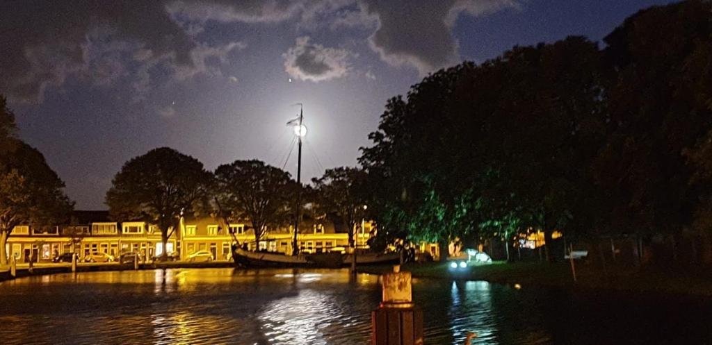 Nachtelijke scène met een verlichte stad aan het water, met een boot op de voorgrond en de maan gloeiend achter wolken.