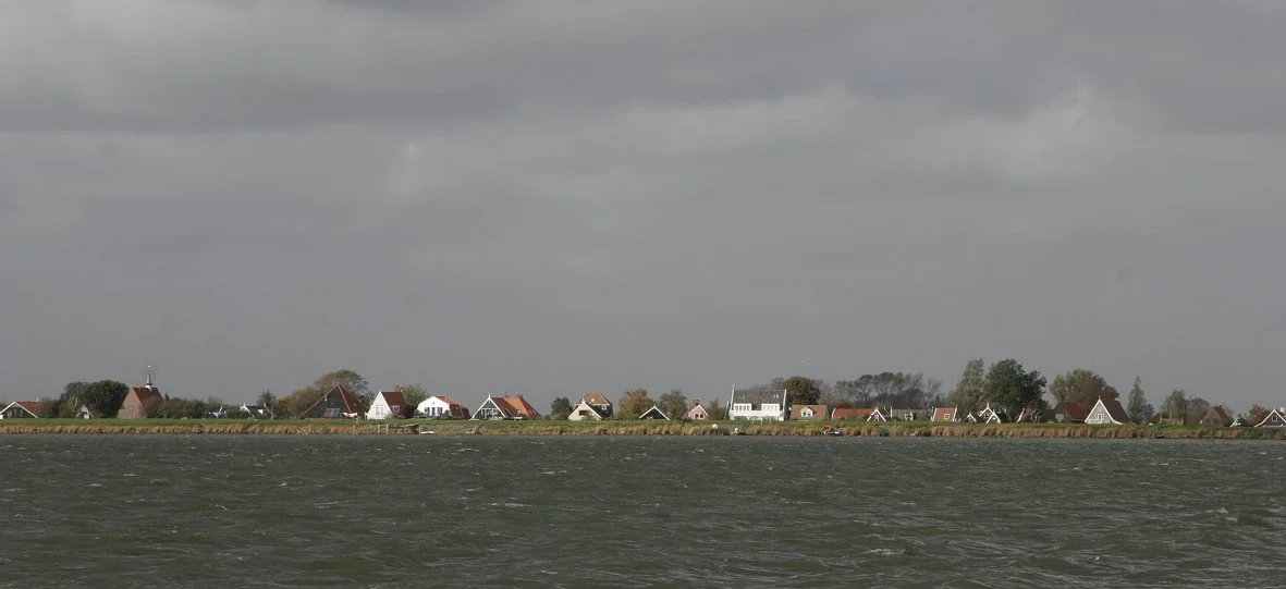 Zicht op een water met een rij huizen aan de kust onder een bewolkte hemel.
