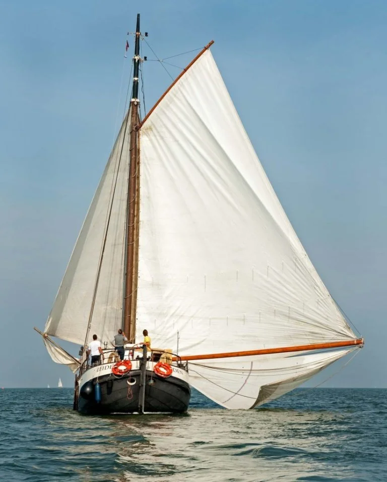 Zeilboot op zee met witte zeilen, bemanningsleden op het dek, heldere blauwe hemel.