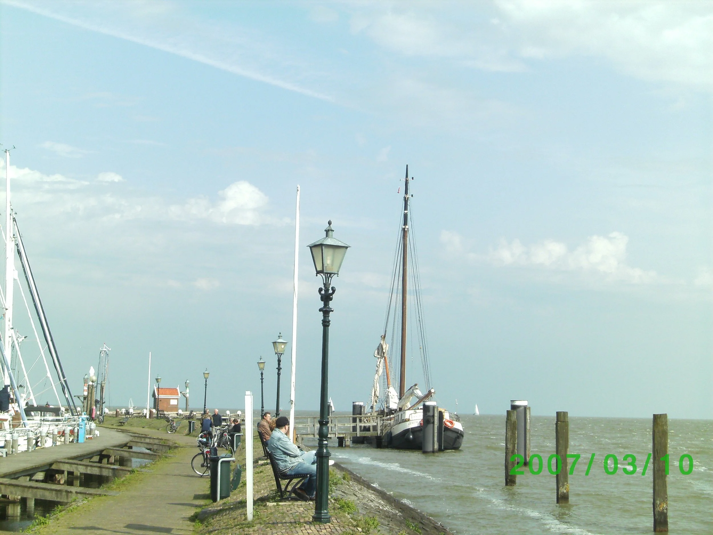 Haven aan een kust met aangemeerde boten, mensen zittend op een bank, en lantaarnpalen langs de kade.