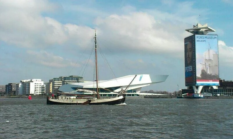 Zicht op een klassieke zeilboot op het IJ in Amsterdam met het EYE Filmmuseum en de A'DAM Toren op de achtergrond, onder een bewolkte lucht.