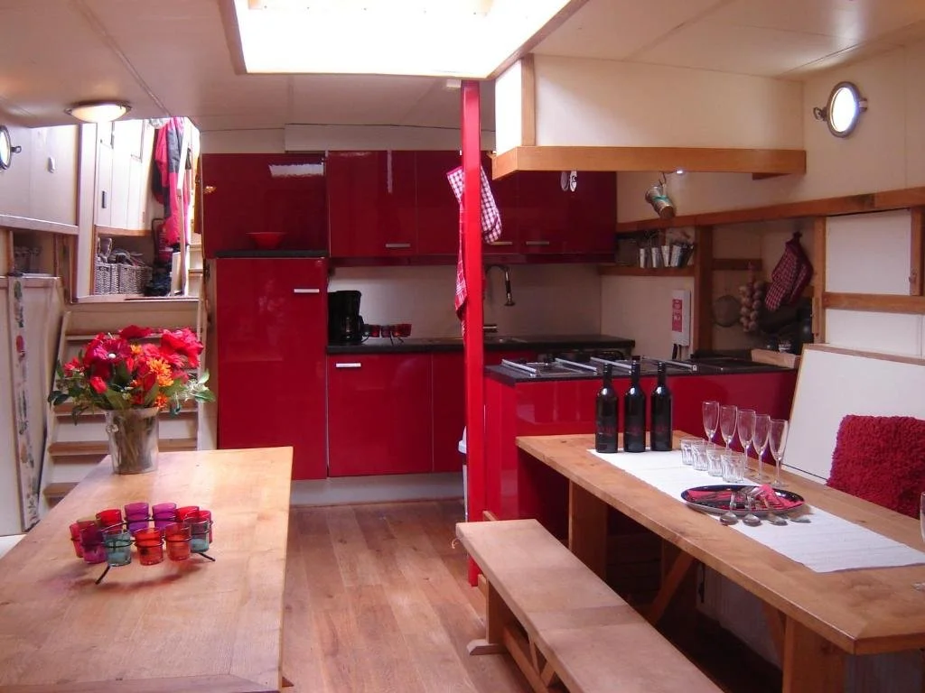 Interieur van een moderne keuken op een woonboot, met een rood keukenblok, houten tafel met flessen wijn en glazen, en een vaas met bloemen.