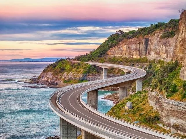 A winding road on a bridge along the coastline with a cliffside and ocean at sunset.