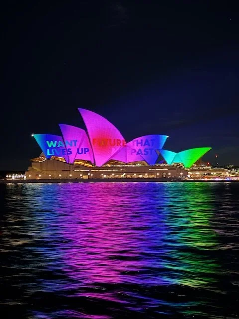 Sydney Opera House with colorful lights projecting the words 'WANT LIVES UP FUTURE THAT PAST' onto its sails, reflected in the water at night.