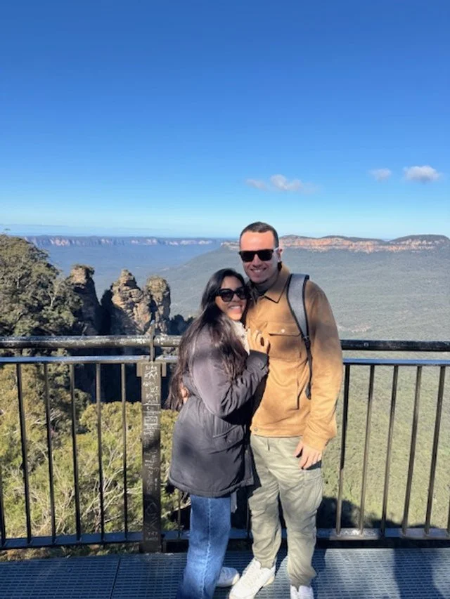 A man and woman standing together on a viewing platform with a scenic canyon, trees, and a clear blue sky in the background.