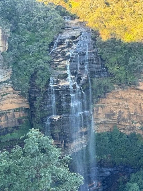 A tall waterfall cascading down a rocky cliff surrounded by green trees, with sunlight shining through the yellow leaves at the top.