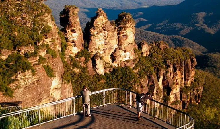 Two people standing on a viewing platform overlooking the Three Sisters rock formation in the Blue Mountains, Australia, with lush green cliffs and forest in the background.