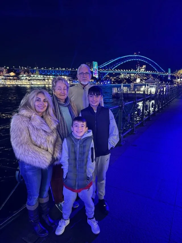 A family of five standing on a waterfront promenade at night with a city skyline and illuminated bridge in the background.