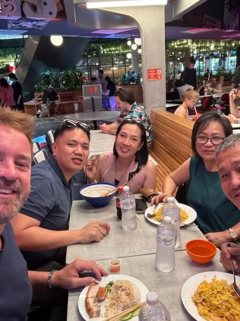 Group of five people sitting at a table in a busy, modern restaurant, enjoying food with plates of rice, curry, and drinks, with other diners and a lit-up interior in the background.