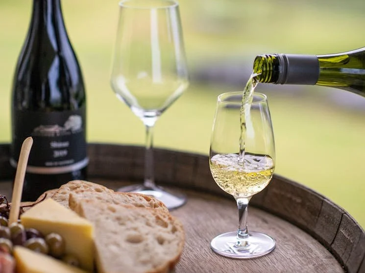 A glass of white wine being poured from a bottle into a wine glass on a wooden tray, with a cheese and bread, and an empty wine glass in the background.
