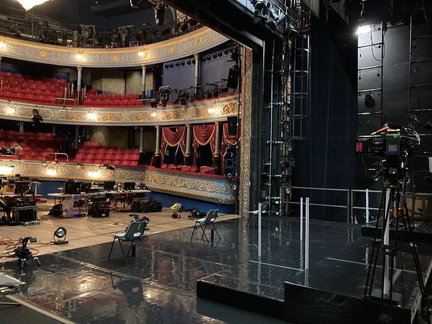 Empty theater stage with chairs and equipment, gold ornate balconies with red seats, black walls and lighting fixtures.