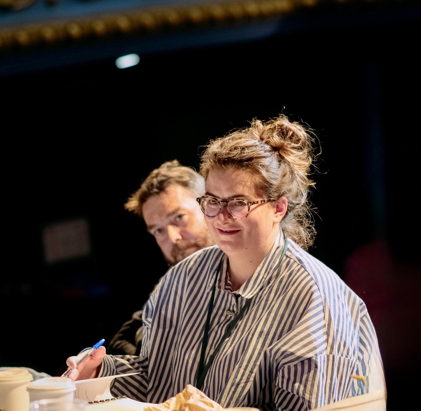 A woman with hair in a messy bun, wearing glasses and a striped shirt, sitting at a table with coffee cups and a notepad, looking slightly to her right with a smile.