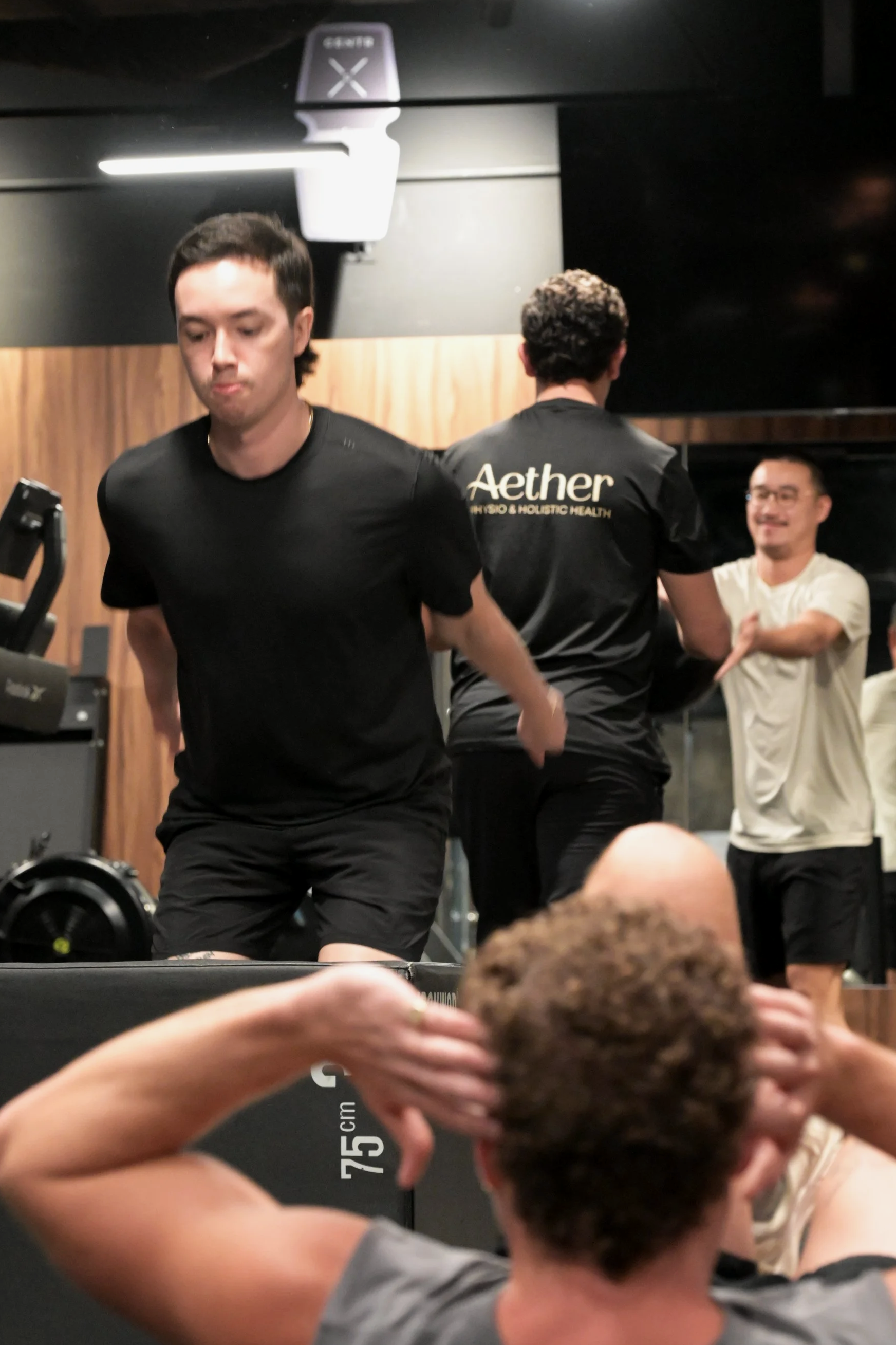 Two men working out in a gym, one facing forward and the other with his back to the camera, with a third person in the background smiling and crossing his arms.
