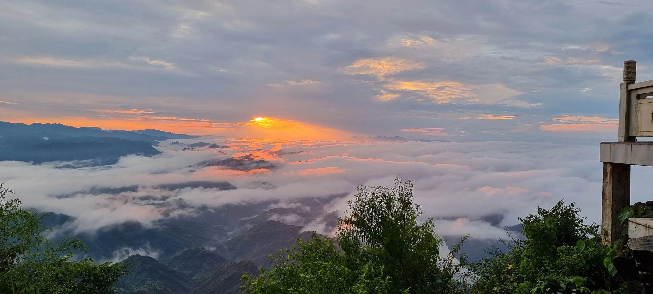 Sunset over mountain range with clouds and fog, some greenery in foreground, part of a balcony or structure on the right side.