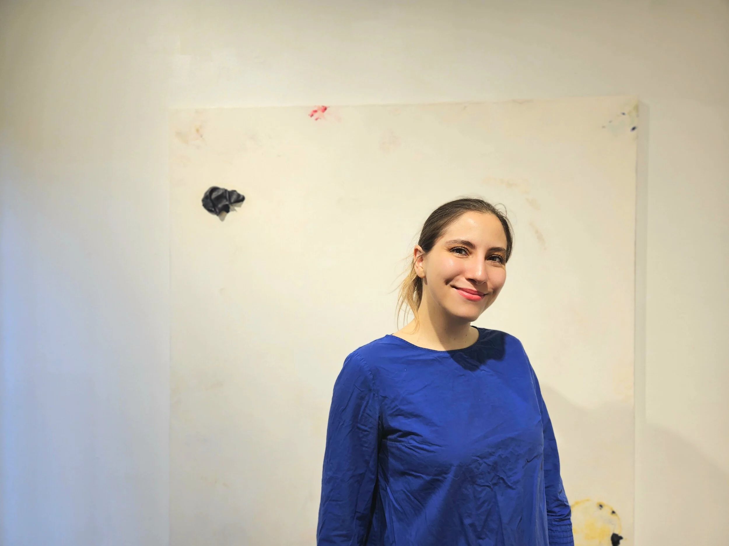 A smiling woman wearing a blue shirt standing in front of a white wall with some paint stains and black tape.