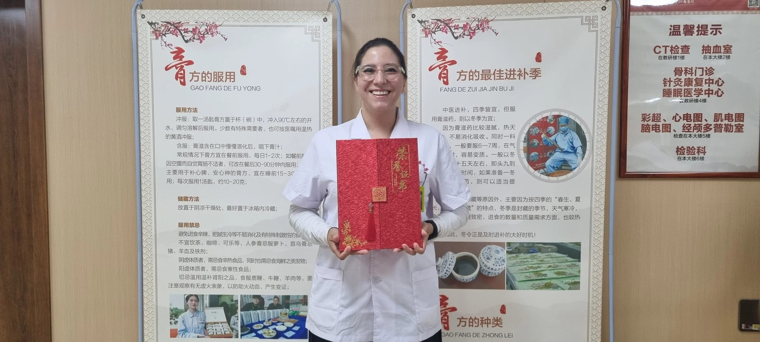A smiling woman in a white medical coat and safety glasses holding a red gift box with gold Chinese characters in front of informational posters in Chinese and images of traditional Chinese medicine and medical workers.