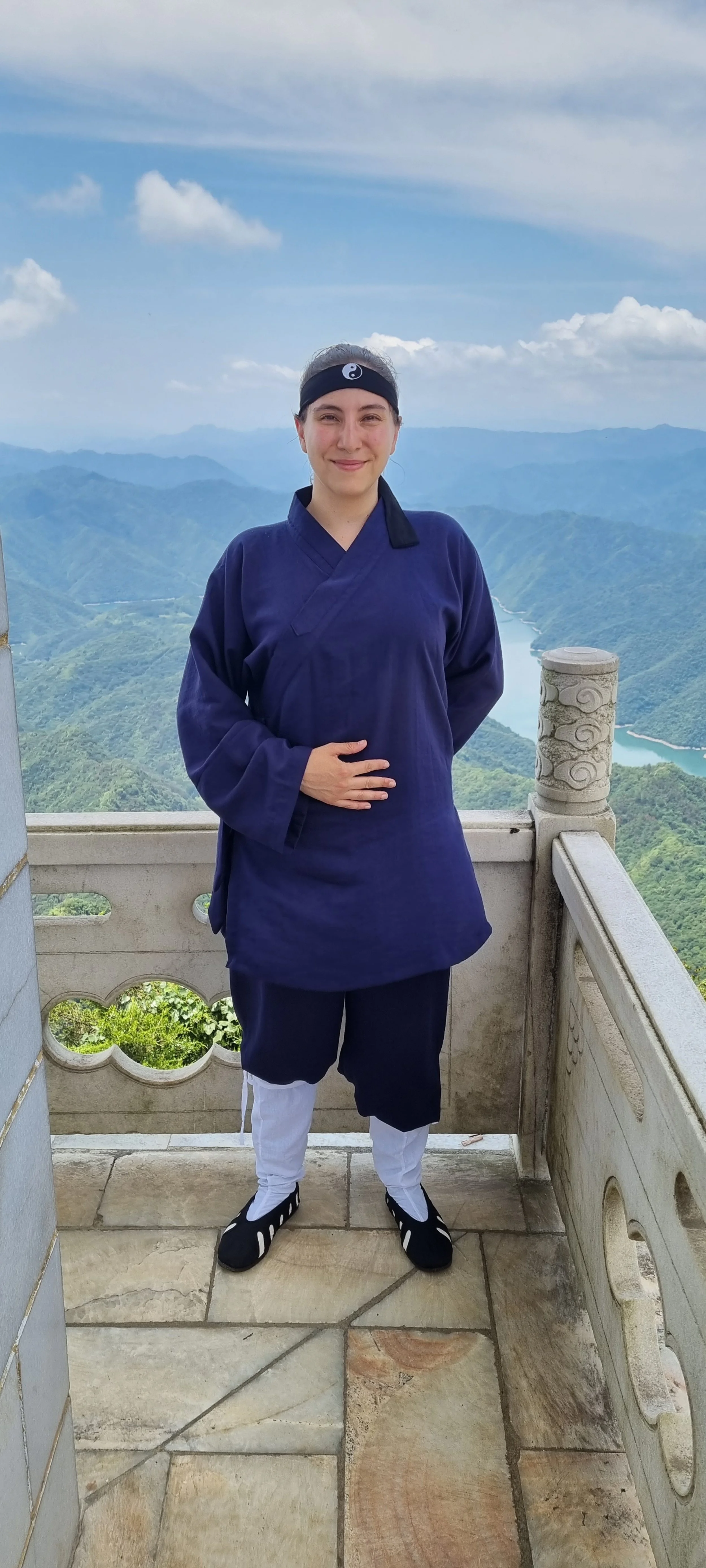 Person in traditional martial arts uniform standing on a mountain viewing platform, with a scenic background of mountains and a river.