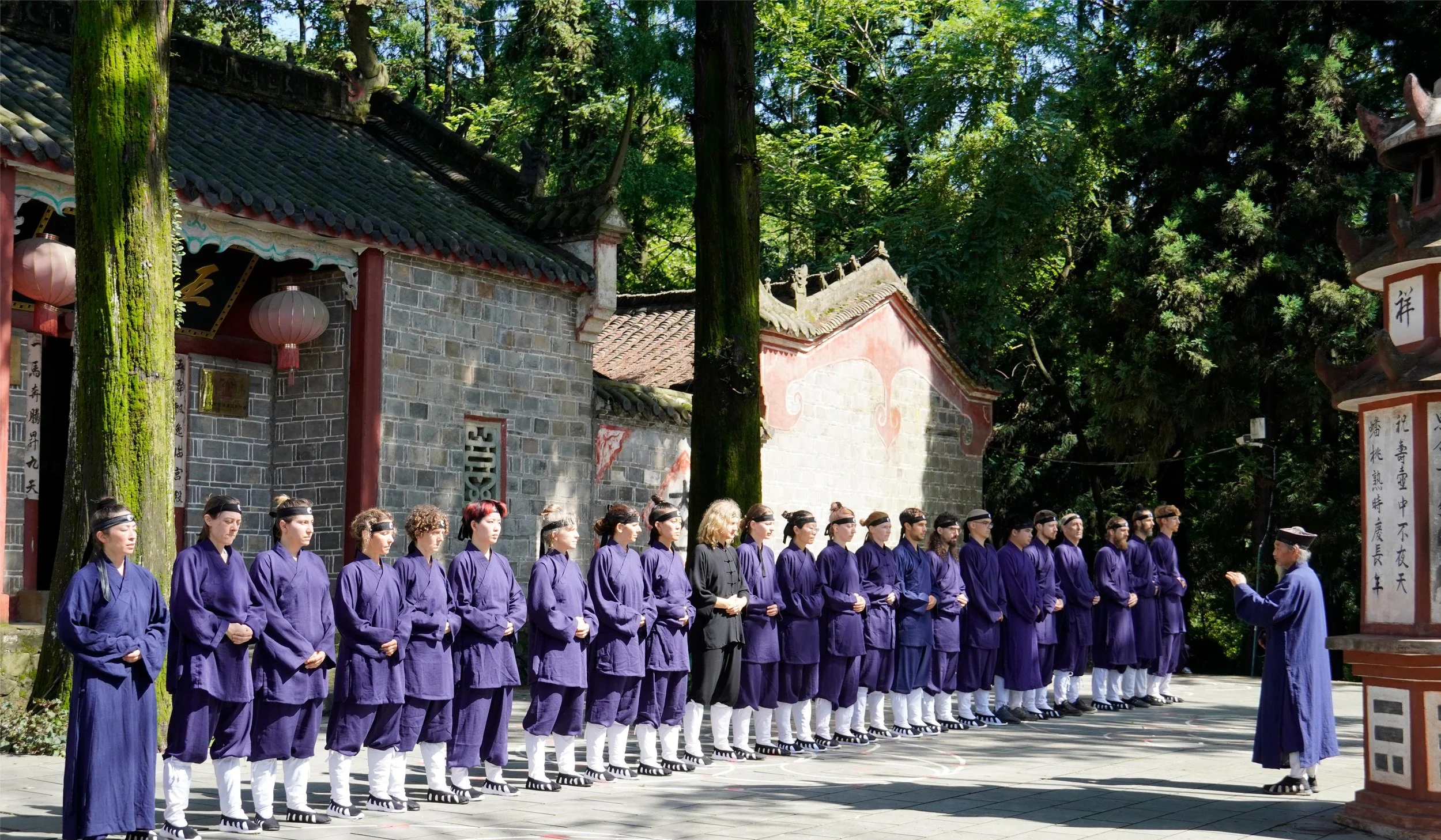 A group of people dressed in traditional Korean clothing standing in line outdoors during daytime, with a person speaking to them, in front of a historical building surrounded by trees.